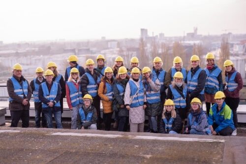 Projekta komanda stāv uz Bērlīnes ūdens apsaimniekošanas uzņēmuma augstākā punkta, visi tērpušies dzeltenās ķiverēs. Starp dalībniekiem ir trīs pārstāvji no Latvijas. Grupas fonā paveras rūpnieciski elementi un apkārtnes ainava