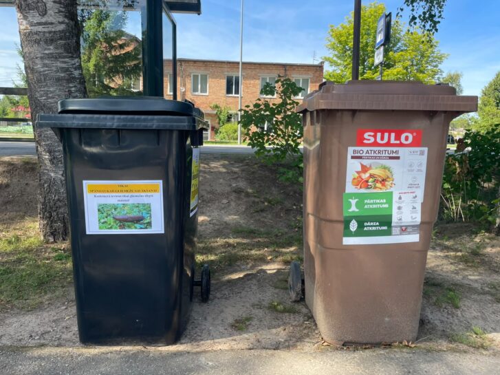 Divas atkritumu tvertnes, melna un brūna, stāv blakus pie koka. Melnajai tvertnei ir uzlīme ar kailgliemeža attēlu un tekstu: "TIKAI SPĀŅU KAILGLIEMEŽU SAVĀKŠANAI. Kaitēkļu savākšanai tikai dārzkopības vajadzībām." Brūnajai tvertnei ir uzlīmes ar tekstu "SULO BIO ATKRITUMI PĀRTIKAS UN DĀRZA", kā arī norādēm "PĀRTIKAS ATKRITUMI" un "DĀRZA ATKRITUMI" ar ikonām. Fonā redzams ceļš un ēka.