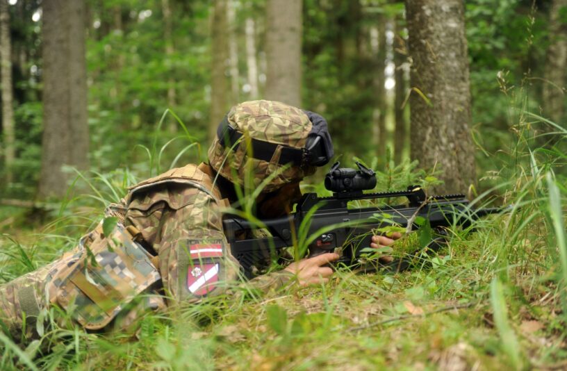 Karavīrs maskēšanās tērpā guļus stāvoklī mežā mērķē ar šauteni. Uz karavīra kreisās rokas redzama uzšuve ar tekstu "LATVIJAS", Latvijas karogu un Zemessardzes ģerboni.