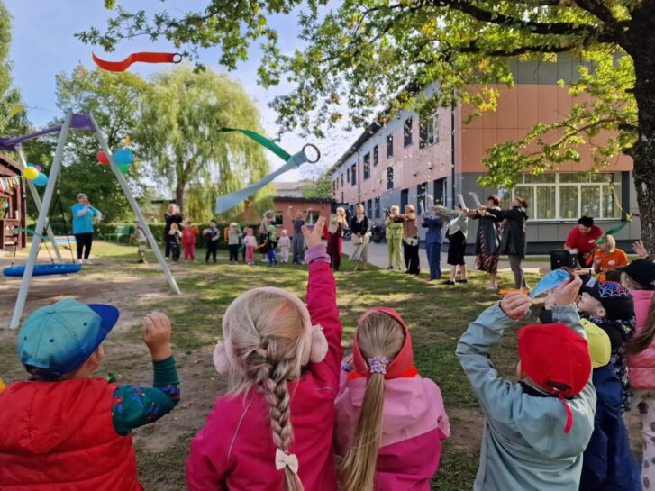 Bērni rotaļu laukumā, no muguras skatoties augšup, kamēr gaisā lido baltas konfeti pārslas un krāsainas lentes. Fonā pie skolas ēkas redzami pieaugušie un citi bērni, kas piedalās āra pasākumā.
