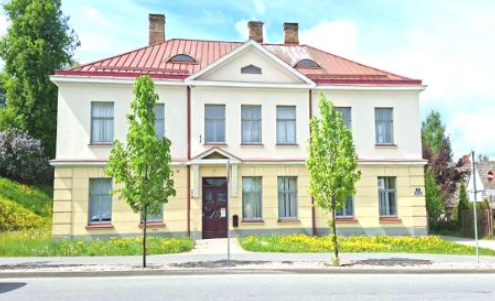 Divstāvu ēka ar sarkanu jumtu un dzeltenīgi baltu fasādi saulainā dienā. Virs ieejas redzams numurs "1", bet uz sānu sienas – zīme "Mākslas skola". Priekšā ir iela ar zālienu, dzelteniem ziediem un diviem jauniem kokiem.