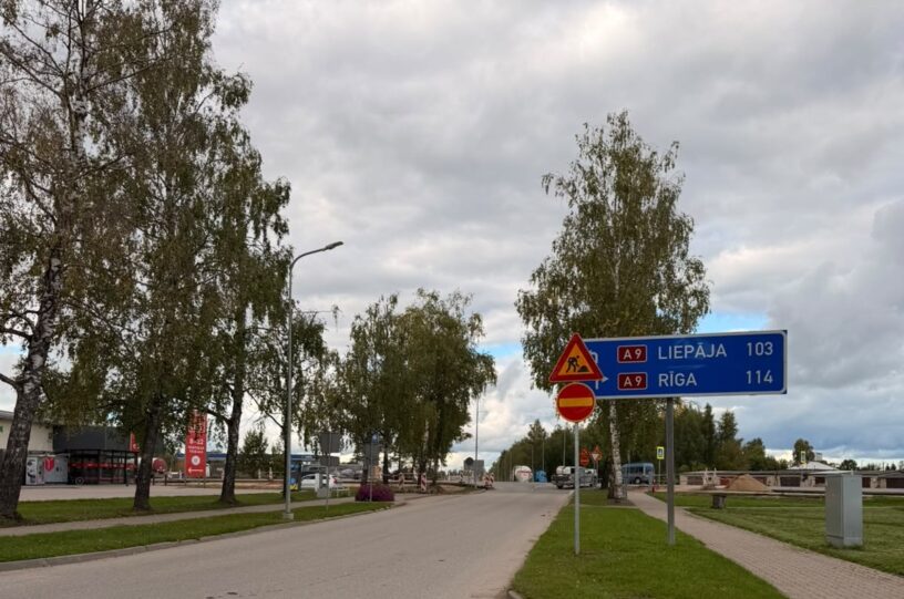 Zilā ceļa zīme norāda uz A9 LIEPĀJA 103 un A9 RĪGA 114. Blakus redzama ceļu darbu un iebraukt aizliegts zīme. Attēlā ir ceļš ar bērziem sānos un apmākušas debesis.