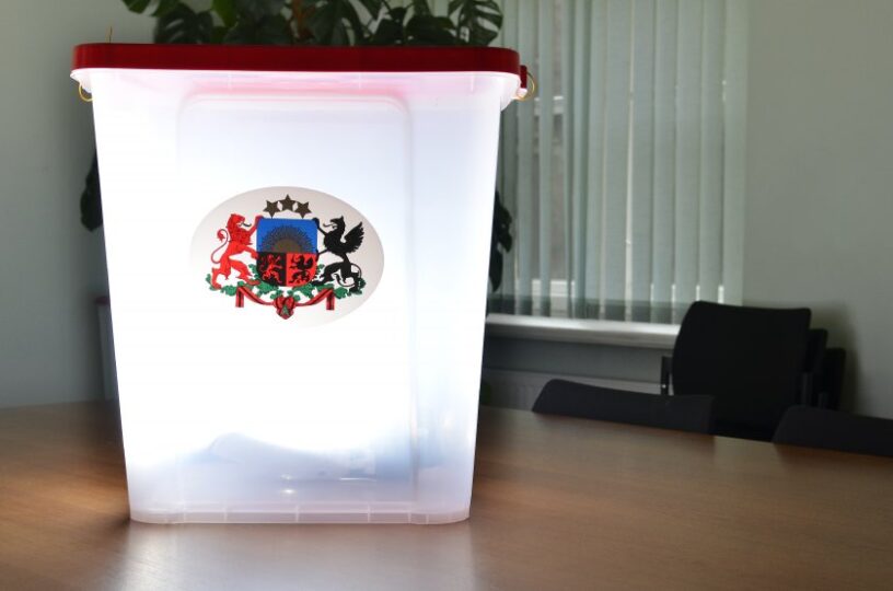 Caurspīdīga plastmasas vēlēšanu urna ar sarkanu vāku, uz kuras ir Latvijas valsts ģerbonis, novietota uz koka galda biroja telpā.