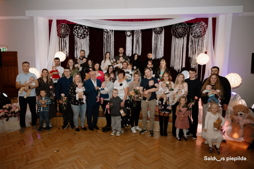 Liela cilvēku grupa, tostarp daudzi pieaugušie, kas tur zīdaiņus, un bērni, pozē grupas fotogrāfijai telpās. Fonā ir tumši sarkani aizkari, balti drapērijas, makramē sapņu ķērāji un spīdošas sfēriskas gaismas. Daži pieaugušie tur rozā rozes. Attēla apakšējā labajā stūrī redzams liels plīša lācis ar rozā lenti un teksts "Saldus piepilda".