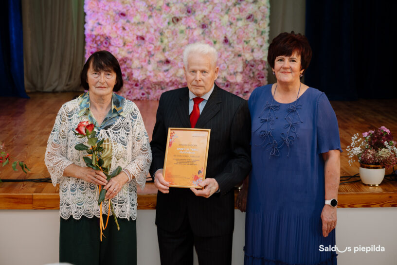 Trīs gados veci cilvēki – divas sievietes un viens vīrietis – stāv uz skatuves ziedu fona priekšā. Kreisajā pusē sieviete tur sarkanu rozi, bet vidū vīrietis tur apbalvojumu ar tekstu "Godīgs darbs Anda un Teiku Rudivicus. 70 gadu kāzu jubilejā.