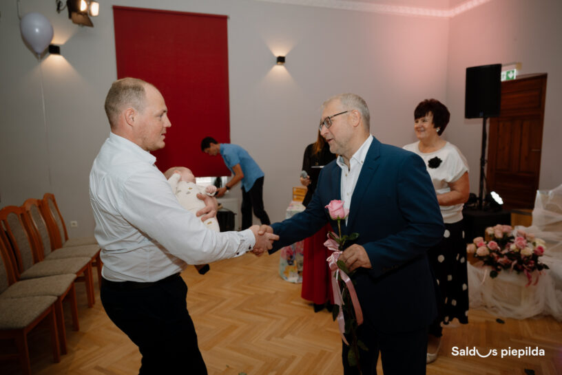 Divi vīrieši, viens turot zīdaini un otrs rozā rozi, rokasspiediena brīdī svinīgā pasākumā. Fonā redzami citi viesi, ziedi un balons. Apakšējā labajā stūrī teksts "Salds piepilda".
