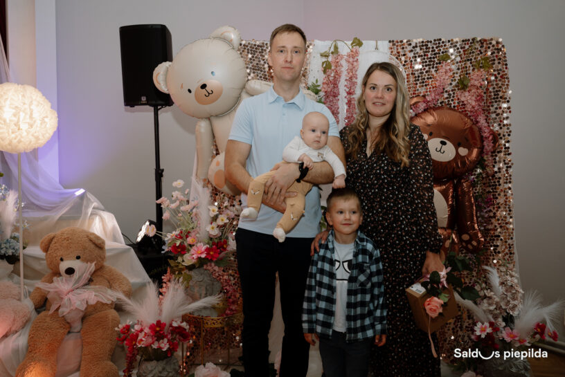 Ģimene – tēvs, māte, zīdainis un mazs zēns – pozē fotogrāfijai ar rotaļu lācīšu baloniem, plīša rotaļu lāci ar rozā svārkiem un ziedu dekorācijām fonā. Tēvs tur rokās zīdaini. Apakšējā labajā stūrī redzams uzraksts "Saldus piepilda".