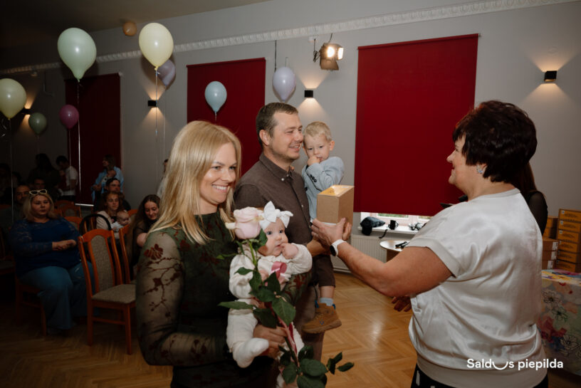 Telpā, kas izrotāta ar baloniem un kurā atrodas viesi, blonda sieviete smaidot tur zīdaini ar rozā rozi. Vīrietis blakus viņai tur mazu zēnu un saņem nelielu dāvanu kastīti no citas sievietes. Attēla apakšējā labajā stūrī ir teksts: "Saldus piepilda".