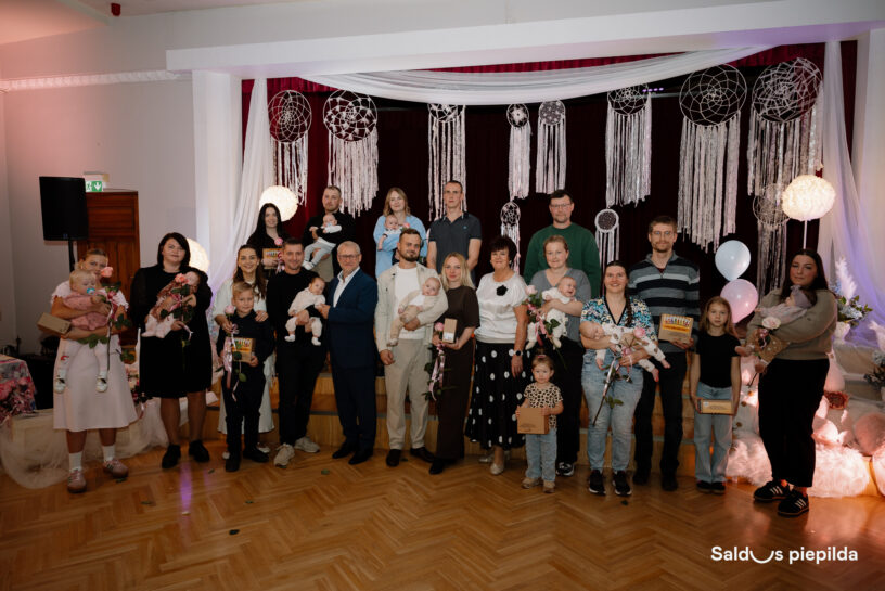 Liela grupa pieaugušo un bērnu, tostarp daudzi zīdaiņi, pozē kopīgai fotogrāfijai. Daudzi pieaugušie tur zīdaiņus, kā arī rozes un apbalvojumus. Fons ir dekorēts ar baltiem makrame sapņu ķērājiem un drapērijām. Apakšējā labajā stūrī redzams teksts "Saldus piepilda".