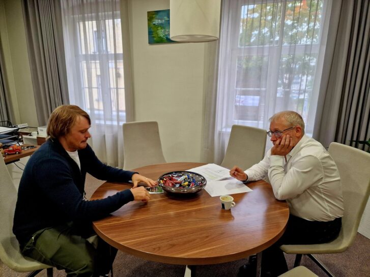 Divi vīrieši sēž pie apaļa koka galda. Jaunākais vīrietis kreisajā pusē, ar gaišiem matiem un ūsām, skatās savā tālrunī. Vecākais vīrietis labajā pusē, ar brillēm, balsta zodu rokā un skatās uz otru vīrieti. Uz galda starp viņiem ir dokumenti, kafijas tase un bļoda ar konfektēm.