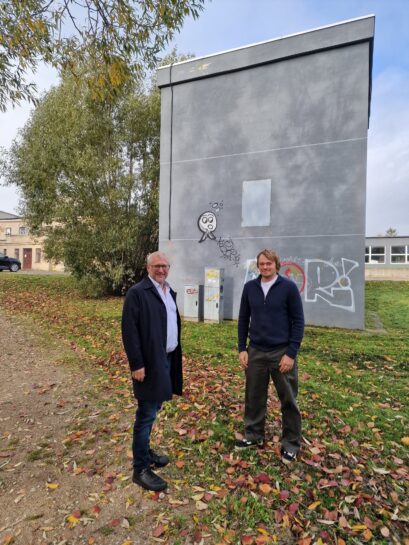 Divi smaidīgi vīrieši stāv laukā pie pelēkas ēkas, kas rotāta ar grafiti zīmējumiem un uzrakstiem. Kreisajā pusē ir vecāks vīrietis brillēs un tumšā mētelī, bet labajā pusē – jaunāks vīrietis tumšā džemperī. Aiz viņiem redzami divi komunālie skapji. Zeme klāta ar kritušām rudens lapām.