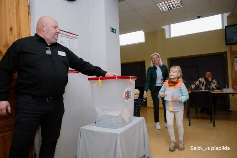 Vīrietis melnā kreklā stāv pie caurspīdīgas vēlēšanu urnas, kurā redzamas daudzas balsu zīmes. Pa labi no viņa sieviete ar diviem bērniem, no kuriem vecākais tur rokā papīra lapu, dodas uz priekšu. un pie galda sēž vēlēšanu darbinieks. Attēla apakšējā labajā stūrī redzama uzraksts "Saldus piepilda".