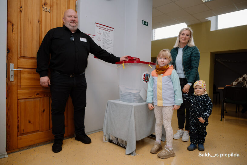 Vīrietis melnā kreklā stāv pie caurspīdīgas vēlēšanu urnas ar Latvijas ģerboni, blakus sieviete ar diviem bērniem.