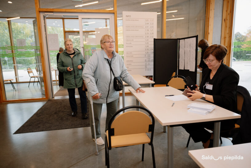Vairākas sievietes reģistrācijas vai informācijas zonā. Priekšplānā sirmgalve ar kruķi, gaišā jakā un brillēm, tuvojas galdam, pie kura sēdoša sieviete melnā apģērbā skatās telefonā un dokumentos. Fonā redzams informatīvs stends ar uzrakstu "INFO" un numuru sarakstu. Attēla apakšējā labajā stūrī lasāms teksts "Saldus piepilda".