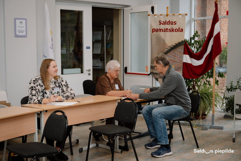 Trīs cilvēki sēž pie galdiem telpā. Pa kreisi jauna sieviete rakstainā blūzē ar vārda zīmīti smaida, skatoties pa labi. Vidū vecāka sieviete brūnā blūzē sarunājas ar vīrieti pelēkā džemperī, kurš noliecis galvu pret galdu. Fonā redzams plakāts ar tekstu "Saldus pamatskola", Latvijas karogs un balts karogs ar zilu logo. Pie sienas ir arī izkārtne ar tekstu "eTwinning School 2023-2024".