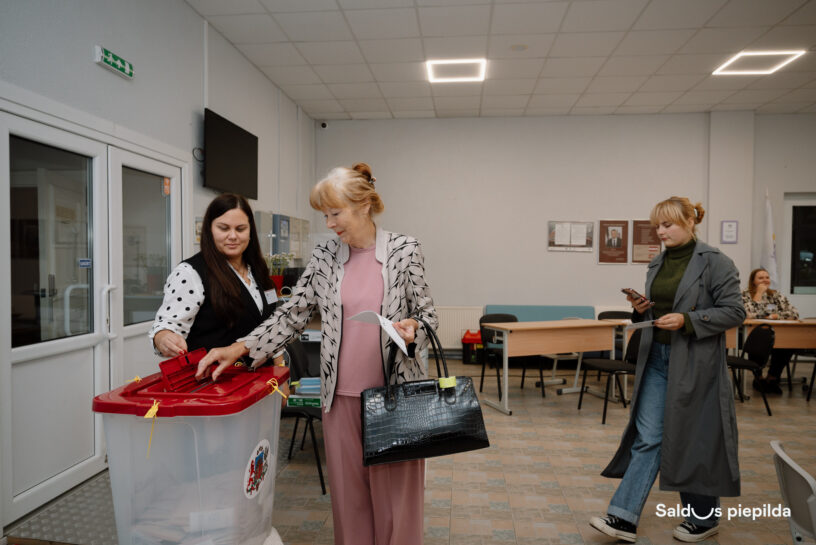 Vēlēšanu iecirknī sieviete ar gaišiem matiem iemet vēlēšanu zīmi caurspīdīgā urnā ar sarkanu vāku un Latvijas ģerboņa attēlu. Viņai blakus stāv vēlēšanu komisijas darbiniece punktotā blūzē. Tālāk labajā pusē redzama jauna sieviete pelēkā mētelī, kas skatās telefonā un tur papīru. Attēla apakšējā labajā stūrī redzams uzraksts "Saldus piepilda".