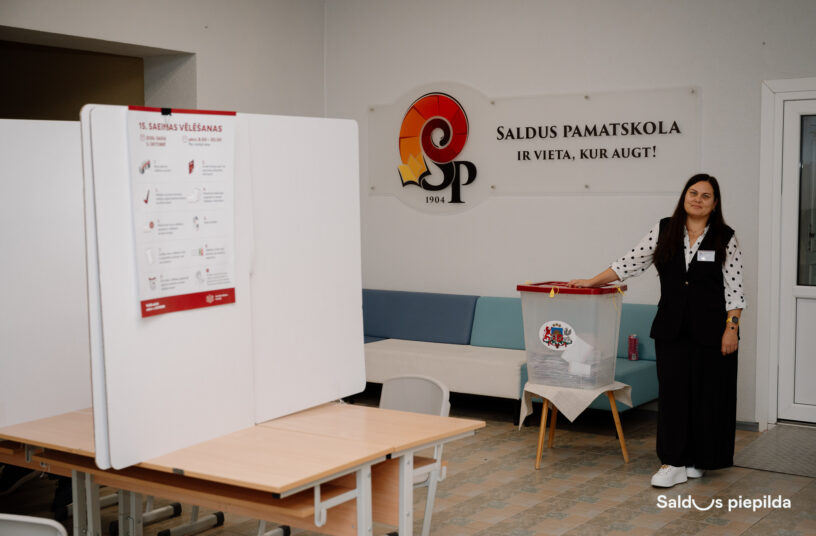 Vēlēšanu iecirknī sieviete ar punktotu kreklu un melnu vesti stāv pie caurspīdīgas balsošanas urnas, uz kuras nolikusi roku. Kreisajā pusē redzama balsošanas kabīne ar plakātu "15. Saeimas vēlēšanas". Fonā pie sienas ir liels uzraksts "SALDUS PAMATSKOLA IR VIETA, KUR AUGT!" ar SP logo un gadskaitli "1904".